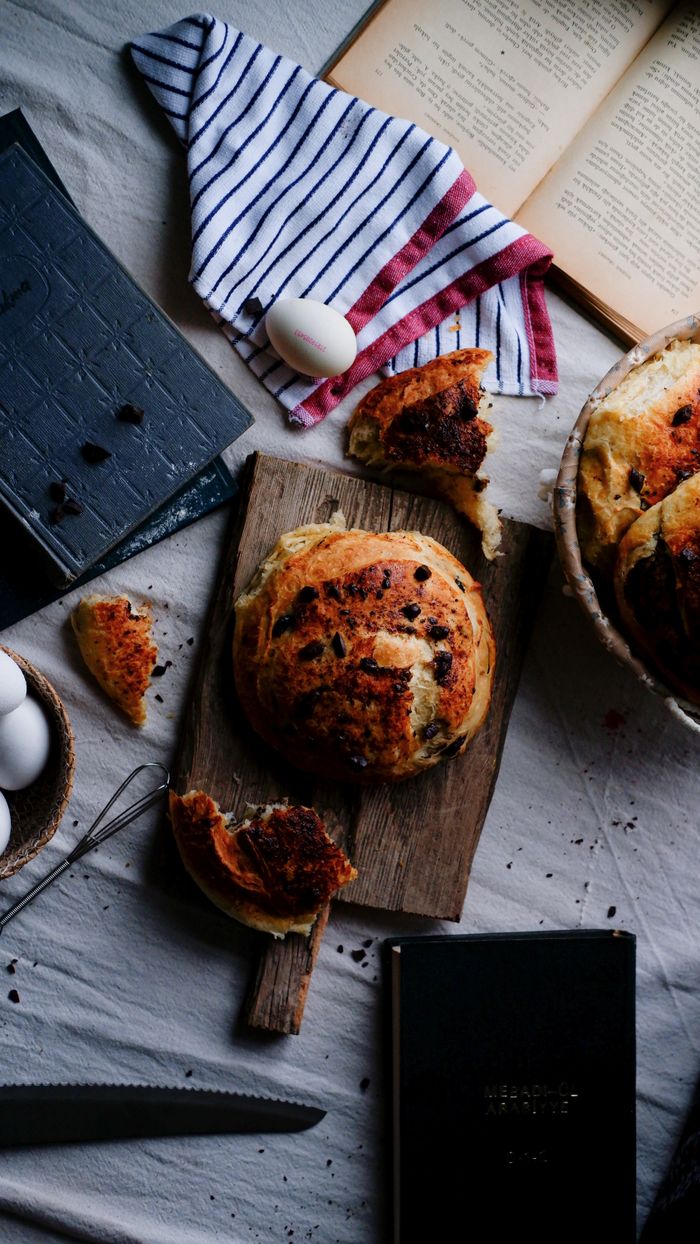 Home A cozy baking scene featuring round bread, eggs, and recipe books on a rustic table setup.